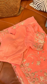 Vasundhara Fashions peach chiffon saree with intricate gold embroidery and sequin detailing on a leather sofa.