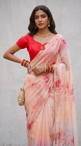 Vasundhara Fashions pink floral organza saree with pearl border paired with a red blouse and gold accessories.